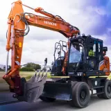 Pomarańczowa koparka Hitachi, gotowa do pracy na złomowisku, pędzi po drodze.