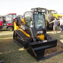 Głównym elementem jest minikoparka gąsienicowa JCB. W tle widać inne maszyny rolnicze i ludzi. Możliwe, że to wystawa, gdzie można sprawdzić jcb polska cennik.
