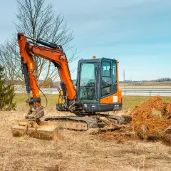 Pomarańczowa minikoparka gotowa do pracy. Trwa wymiana gąsienicy, maszyna stoi na nierównym, trawiastym terenie obok usypanej ziemi.