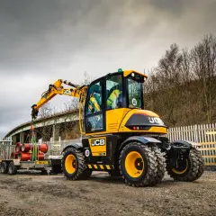 Żółta koparka JCB Hydradig, ważąca ok. 11 ton, ładuje rury na przyczepę.