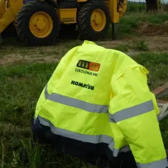 Kamizelka z logo Komatsu i napisem "Szkolenia MM" leży na krześle obok koparki. Idealna na testy na koparki jednonaczyniowe.