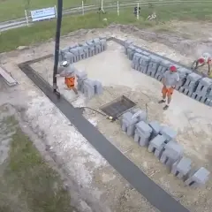 Pracownicy wylewają beton na fundamenty. Widać koparkę, betoniarkę i sterty pustaków.