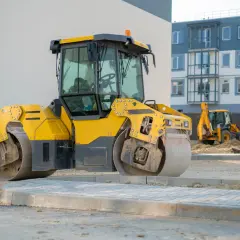 Żółty walec drogowy przygotowuje teren pod budowę. W tle widać koparkę i nowe budynki.