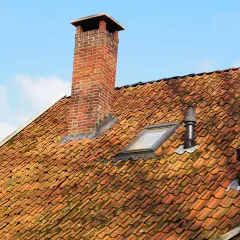 Ceglany komin i dwa czarne kominki wentylacyjne na pomarańczowym dachu.