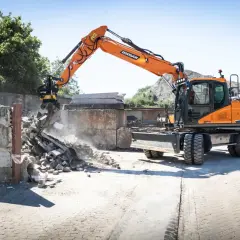 Pomarańczowa koparka Doosan DX150W pracuje na placu budowy w Poznaniu, rozbierając betonowe ściany.