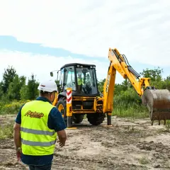 Mężczyzna w kasku i kamizelce odblaskowej obserwuje koparko-ładowarkę, przygotowując się do egzaminu na operatora.