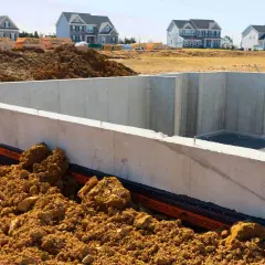 Fundament betonowy w budowie, otoczony ziemią. W tle nowe domy. Zastanawiasz się, ile kosztuje beton na fundamenty?