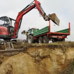 Czerwona koparka do stawu pracuje, zasypując ziemią przyczepę traktora.