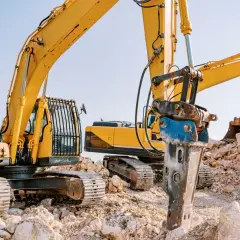 Żółte koparki na placu budowy, jedna z nich wyposażona w młot udarowy hydrauliczny do kruszenia kamieni.