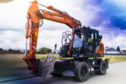 Pomarańczowa koparka Hitachi, gotowa do pracy na złomowisku, pędzi po drodze.