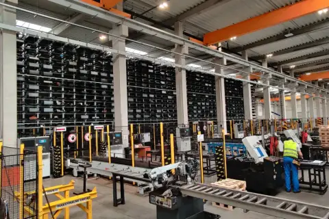 Magazyn z regałami pełnymi materiałów, w tym różnych gatunków stali trudnościeralnej. Pracownik w kamizelce odblaskowej przy maszynie.