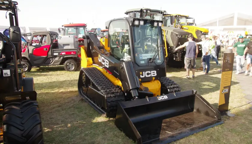 Głównym elementem jest minikoparka gąsienicowa JCB. W tle widać inne maszyny rolnicze i ludzi. Możliwe, że to wystawa, gdzie można sprawdzić jcb polska cennik.