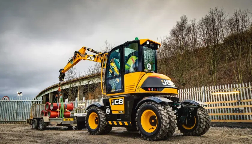 Żółta koparka JCB Hydradig, ważąca ok. 11 ton, ładuje rury na przyczepę.