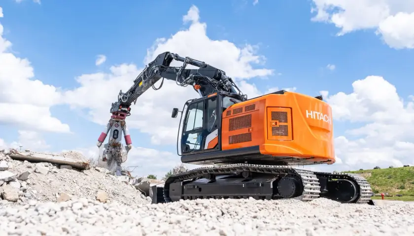 Pomarańczowa koparka do rozbiórek Hitachi z chwytakiem pracuje na gruzowisku pod błękitnym niebem z chmurami.