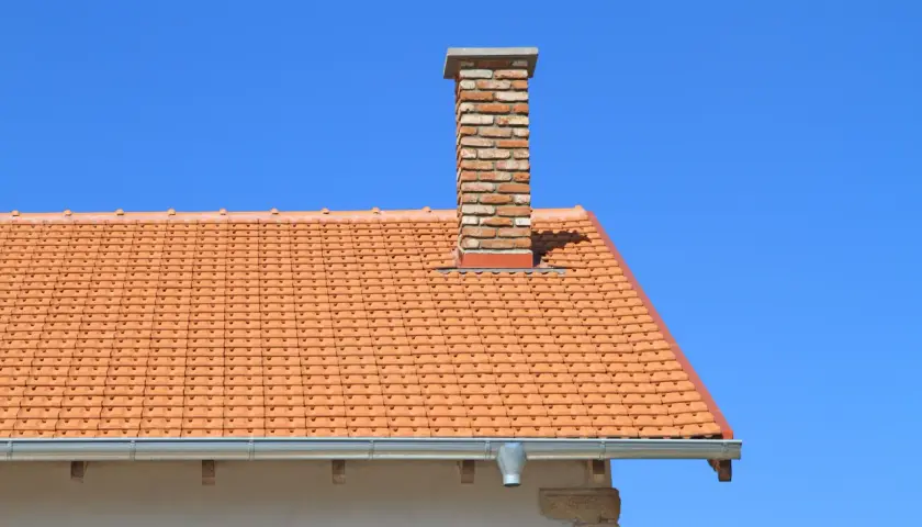 Ceglany komin na dachu z pomarańczowej dachówki. Zastanawiasz się, ile kosztuje budowa komina?