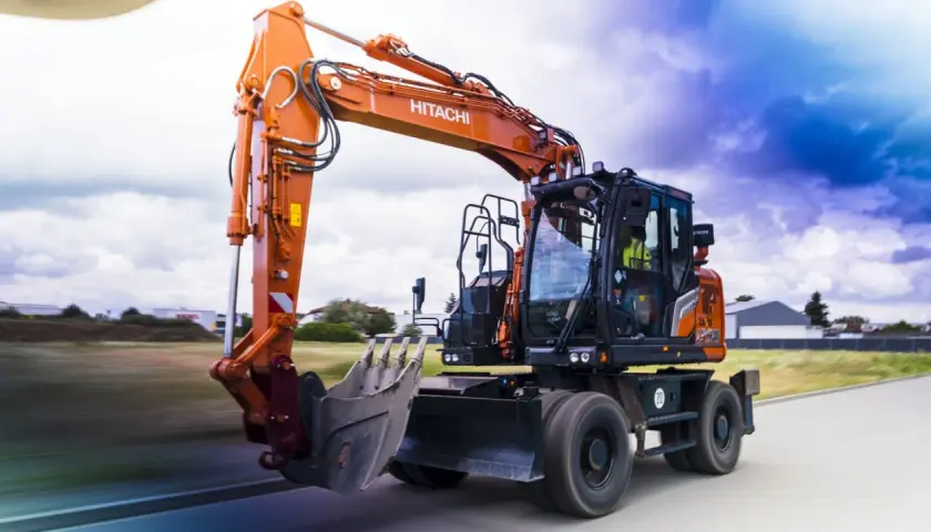 Pomarańczowa koparka Hitachi, gotowa do pracy na złomowisku, pędzi po drodze.