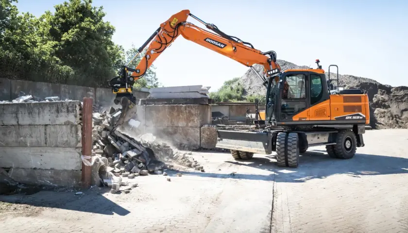 Pomarańczowa koparka Doosan DX150W pracuje na placu budowy w Poznaniu, rozbierając betonowe ściany.