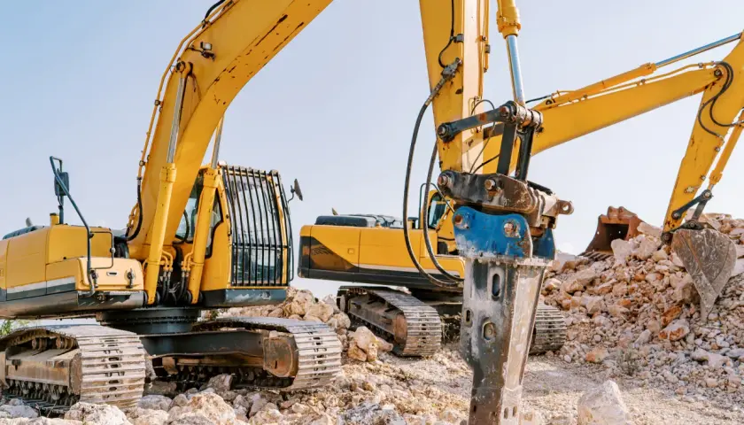 Żółte koparki na placu budowy, jedna z nich wyposażona w młot udarowy hydrauliczny do kruszenia kamieni.