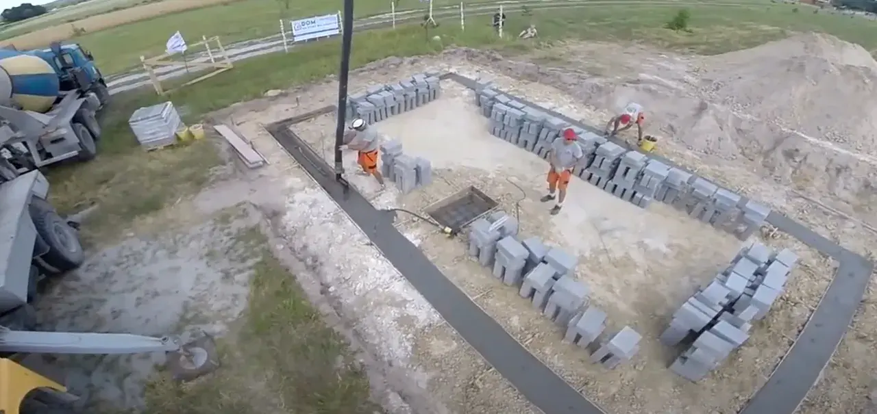 Pracownicy wylewają beton na fundamenty. Widać koparkę, betoniarkę i sterty pustaków.