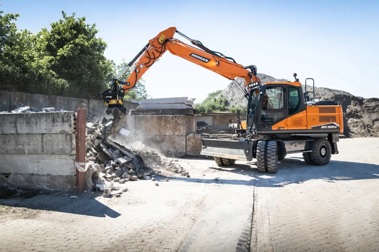 Pomarańczowa koparka Doosan DX150W pracuje na placu budowy w Poznaniu, rozbierając betonowe ściany.