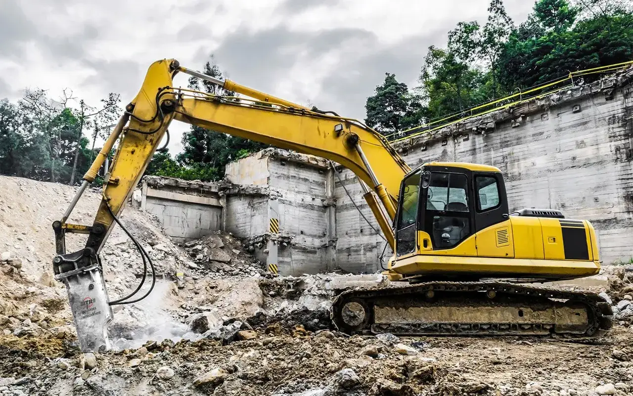 Żółta koparka z młotem udarowym hydraulicznym kruszy beton na placu budowy.