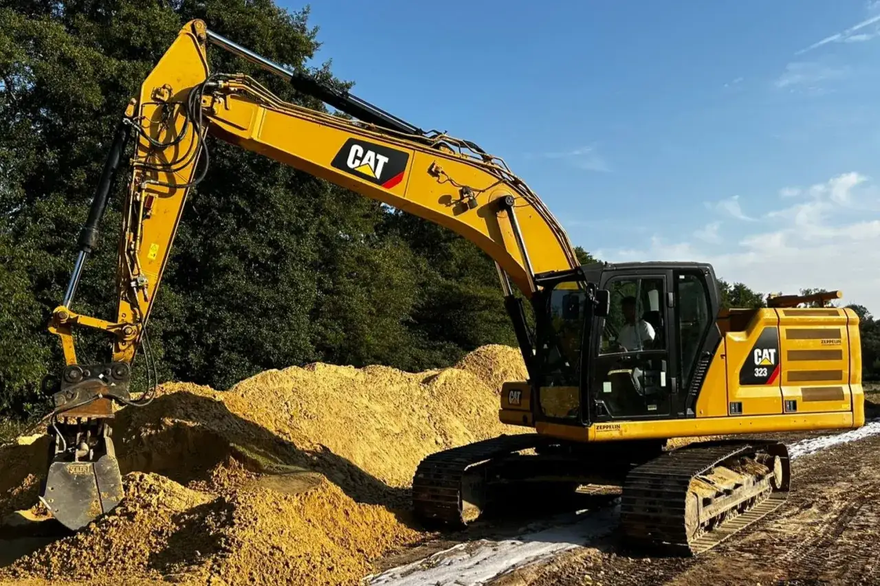 Żółta koparka gąsienicowa CAT 323 pracuje na budowie, przesuwając piasek. Warto wiedzieć, ile kosztuje godzina pracy koparki gąsienicowej.