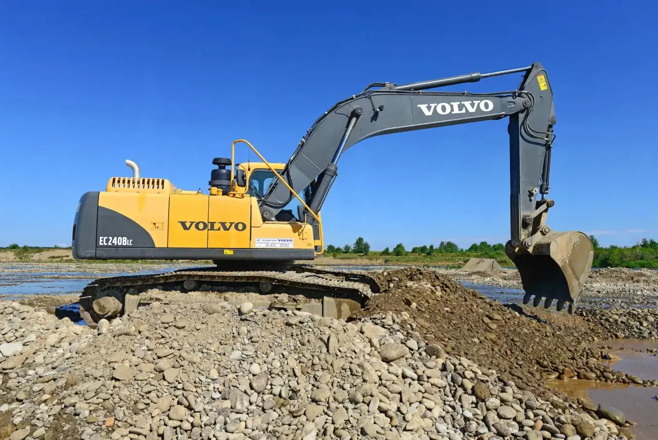 Żółta koparko-ładowarka Volvo pracuje nad zleceniem na koparko ładowarkę, przesuwając kamienie i ziemię wzdłuż rzeki.