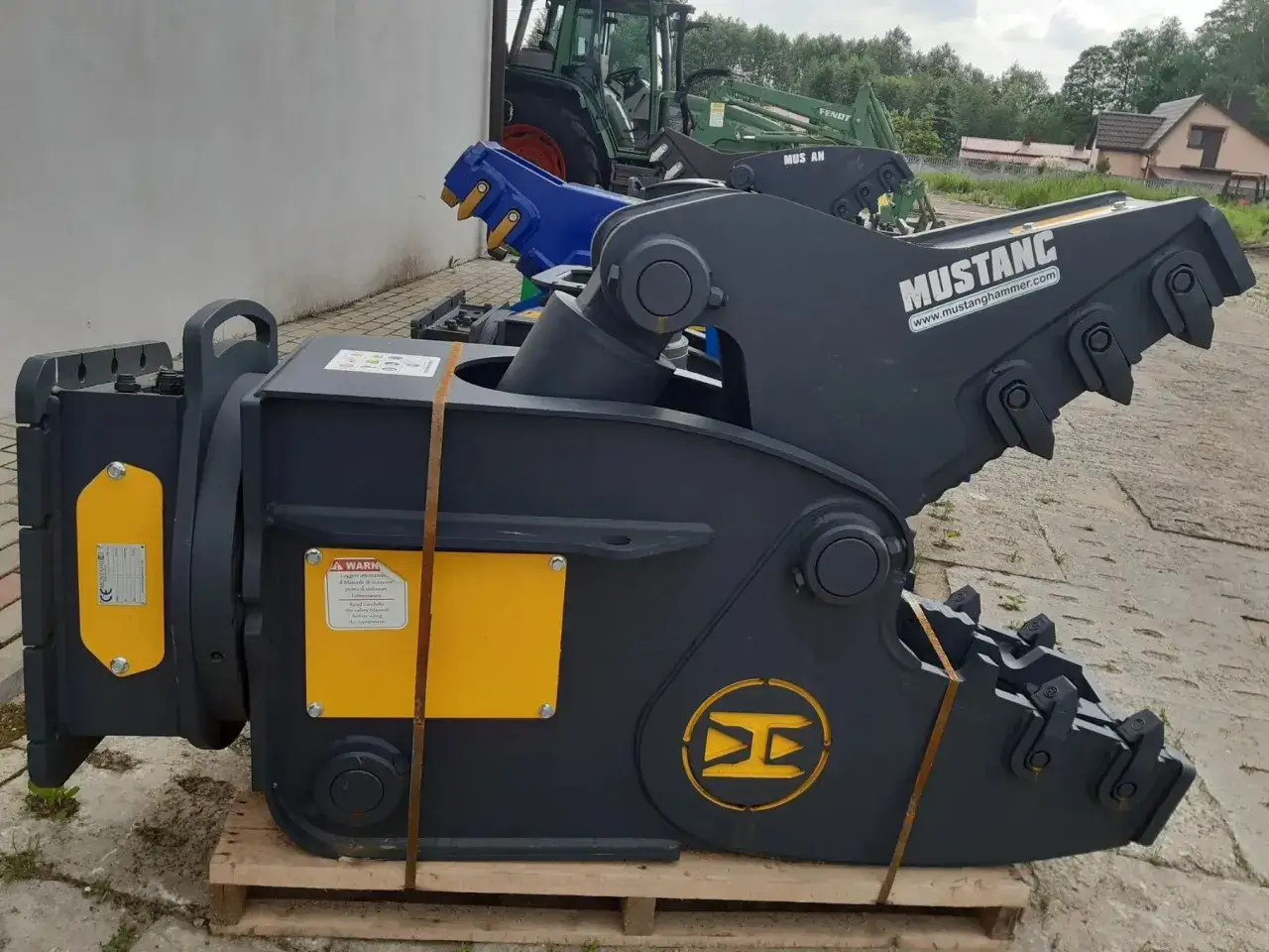 Solidne nożyce hydrauliczne do koparki Mustang, gotowe do pracy. W tle zielony traktor.