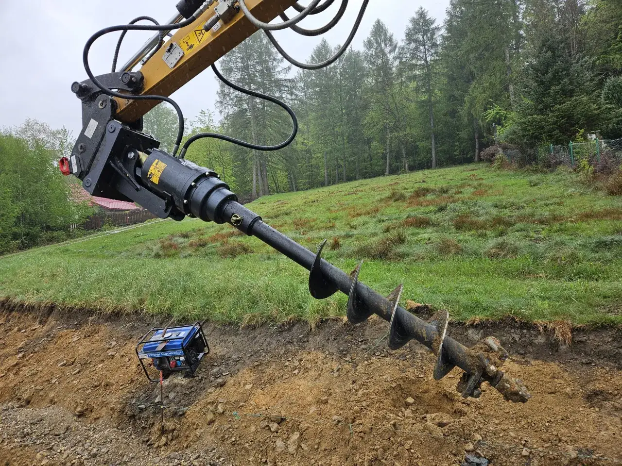 Mocny młot udarowy hydrauliczny z augerem przygotowuje teren pod budowę, wiercąc w ziemi. W tle las i zielona trawa.