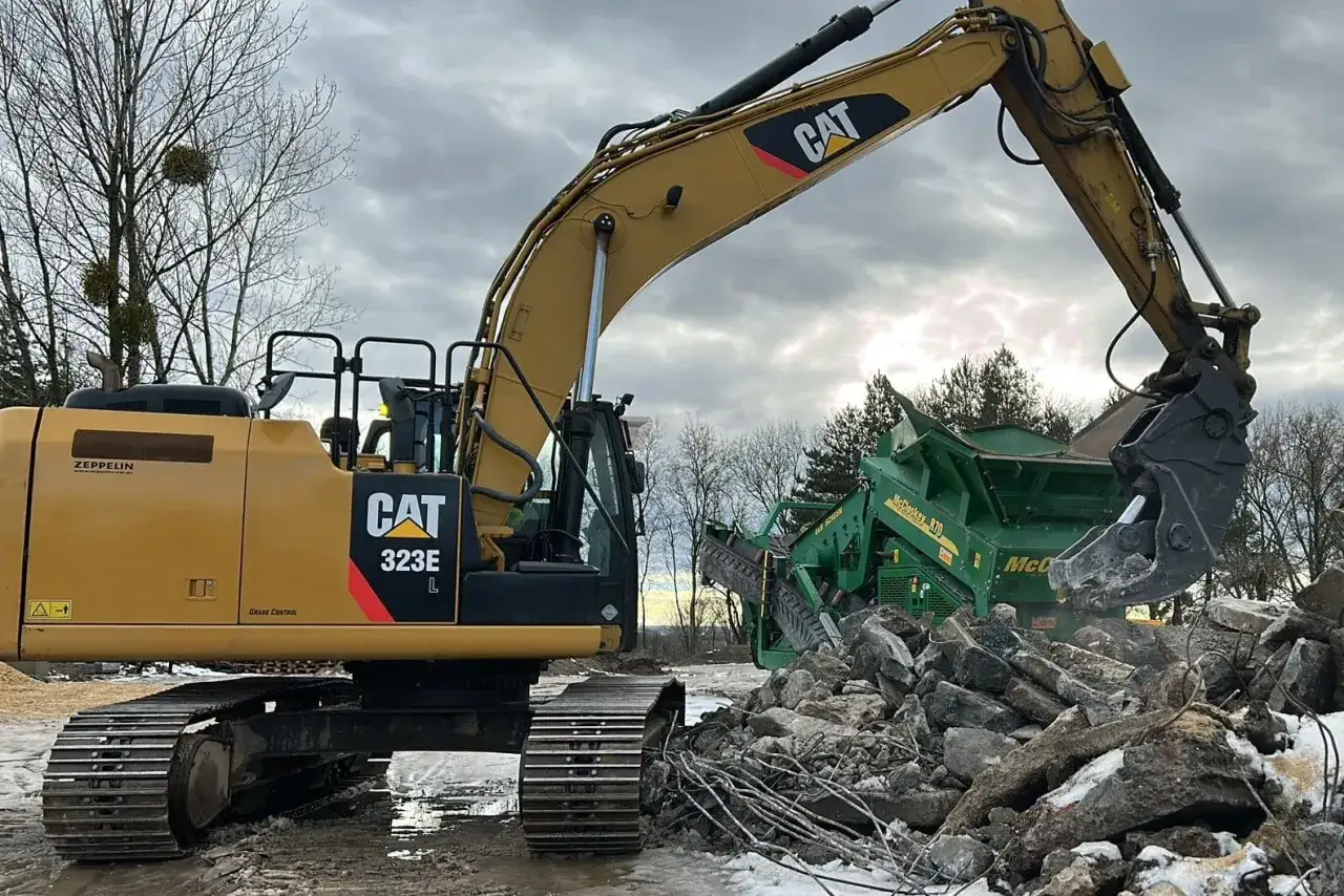 Żółta koparka CAT 323E pracuje przy kruszarce. Zastanawiasz się, ile kosztuje godzina pracy koparki gąsienicowej?