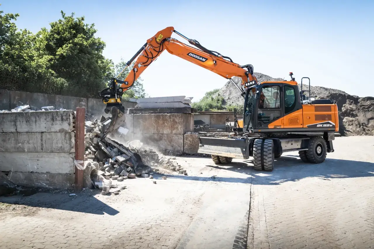 Pomarańczowa koparka Doosan DX150W pracuje na placu budowy w Poznaniu, rozbierając betonowe elementy.
