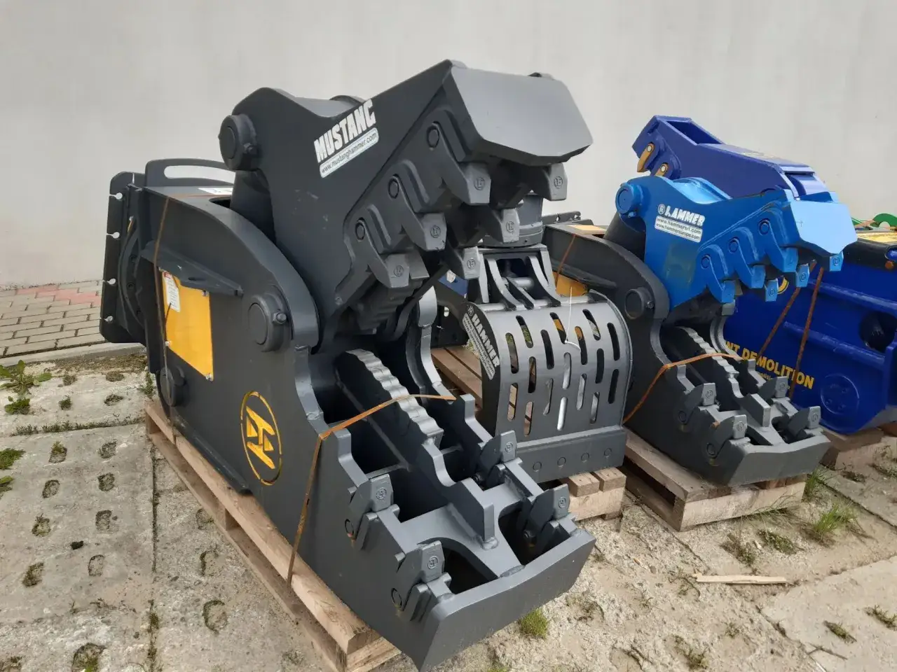 Dwa potężne nożyce hydrauliczne do koparki, jeden szary Mustang, drugi niebieski Hammer, gotowe do pracy.