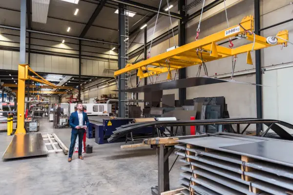 Pracownik w garniturze analizuje dokumentację, zastanawiając się, ile kosztuje kurs na suwnicę, podczas gdy maszyna podnosi ciężki ładunek.